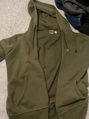 Olivgrön hoodie med dragkedja - Snygg olivgrön hoodie med dragkedja och huva. Tröjan har fickor framtill och justerbara snören i huvan. Tillverkad i mjukt material, perfekt för en avslappnad stil. Liten broderad logga på bröstet för en clean look.