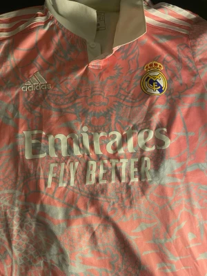 Real Madrid rosa matchtröja Adidas - Säljer en rosa Real Madrid matchtröja från Adidas med ljusblått drakmönster. Tröjan har klubbmärke och Adidas-logga på bröstet samt Emirates Fly Better-tryck på framsidan. Perfekt för fotbollsträning eller att bära som supporter.