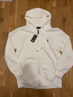 Vit hoodie från Polo Ralph Lauren - Snygg vit hoodie från Polo Ralph Lauren med dragkedja framtill och klassisk svart logga broderad på bröstet. Hoodien har huva med dragsko, ribbade muddar och kängurufickor. Tillverkad i mjukt bomullsmaterial för en skön känsla.