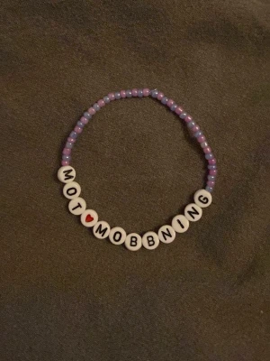 Pärlarmband med text mot mobbning - Säljer ett handgjort armband med lila och vita pärlor samt bokstavspärlor som stavar 'MOT MOBBNING' med ett rött hjärta. Armbandet är elastiskt och har en ungdomlig och personlig stil. Perfekt för dig som vill visa ditt ställningstagande.