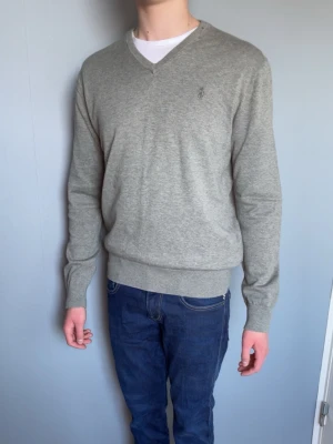 Grå V-ringad tröja från Ralph Lauren - Snygg grå V-ringad tröja från Ralph Lauren med diskret broderad logga på bröstet. Tröjan har långa ärmar, ribbade muddar och är tillverkad i mjukt material som känns skönt mot huden. Perfekt att bära över en t-shirt.