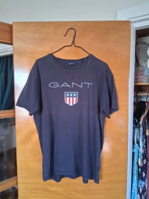 Svart GANT t-shirt med logga - Snygg svart t-shirt från GANT med klassisk logga och sköld på bröstet. T-shirten har rund halsringning och korta ärmar. Perfekt för en stilren och avslappnad look! 😃 Finns ett nindre hål framtill, kan lagas innan frakt, om så önskas!