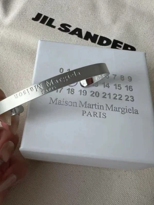 Maison Margiela silverfärgat armband - Stilrent armband i silverfärgad metall från Maison Martin Margiela Paris. Armbandet är stelt och har en graverad logotyp längs utsidan. Perfekt accessoar för dig som gillar minimalistisk och modern design. Använd sparsamt i några månader och är i toppskick!