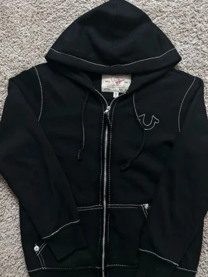  True Religion  - Svart hoodie med dragkedja från True Religion. Snygga vita kontrastsömmar, logga broderad på bröstet och stort tryck med text och grafik på ryggen. Huva med dragsko och ribbade muddar. Perfekt för en streetwear-look.