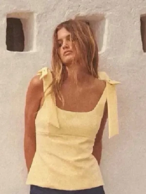 Gul linnetopp med knytband - Snygg ljusgul linnetopp med breda axelband som knyts i rosetter. Toppen har en rak och något croppad passform, perfekt för varma dagar. Materialet ser ut att vara linne och ger en fräsch och somrig känsla.