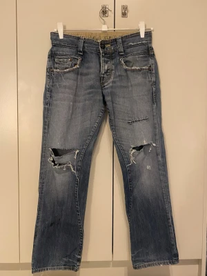 Blå slitna jeans från G-Star RAW - Säljer ett par blå jeans från G-Star RAW med tydliga slitningar och hål på båda benen. Jeansen har fem fickor, raka ben och klassisk knappgylf. Materialet är denim i bomull och de har en cool, avslappnad look. Har lite fläckar på ena benet men syns inte jättetydligt 