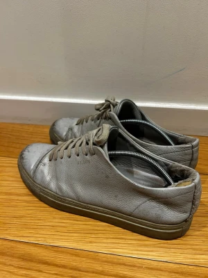 Gråa sneakers från ARIGATO i läder *43* - Snygga gråa sneakers från ARIGATO i mjukt läder med klassisk låg profil och matchande grå snörning. Skorna har en stilren design med diskret logga på sidan och en slitstark gummisula. Perfekta för en clean och minimalistisk look.