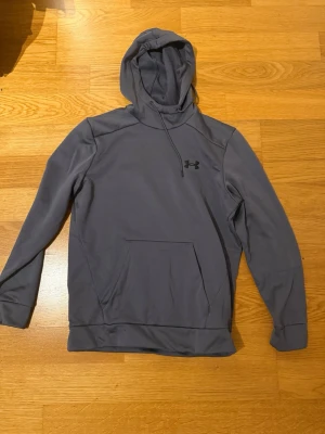 Grå hoodie från Under Armour - Snygg grå hoodie från Under Armour med klassisk känguruficka och justerbar huva. Tröjan har en stilren design med logga på bröstet och är tillverkad i ett mjukt, sportigt material som passar perfekt till chill eller träning.
