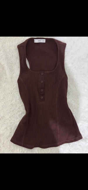 Brun ribbad linnetopp från Mango - Snygg brun ribbad linnetopp från Mango med rund halsringning och knappdetaljer framtill. Toppen är ärmlös och har en figurnära passform som framhäver siluetten. Perfekt att styla med jeans eller kjol för en trendig look. Aldrig använd🤎🤎
