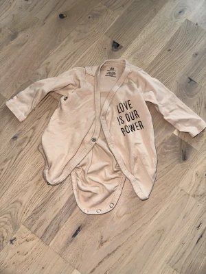 Beige omlottbody från H&M - Säljer en beige långärmad omlottbody från H&M i mjuk bomull. Tryckknappar längs sidan och i grenen för enkel påklädning. Texten 'LOVE IS OUR POWER' tryckt framtill ger en cool touch. Perfekt för små barn som vill ha något bekvämt och stilrent.