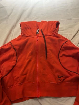 Röd Nike Air hoodie med dragkedja - Snygg röd hoodie från Nike Air med hel dragkedja och svart logga på framsidan. Hoodien har huva med svarta snören och coola svarta detaljer längs ärmarna. Croppad modell som passar perfekt till streetwear-stilen.