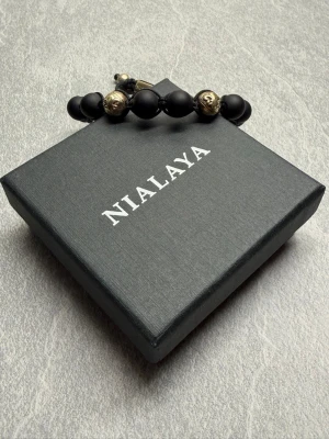 Svart pärlarmband från Nialaya - Stilrent armband från Nialaya med mattsvarta pärlor och detaljer i guldton. Armbandet har ett justerbart snöre och coola guldfärgade pärlor med mönster som ger en unik touch. Perfekt accessoar för dig som gillar en modern och edgy stil.