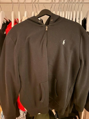 Svart hoodie från Polo Ralph Lauren - Klassisk svart hoodie med dragkedja från Polo Ralph Lauren. Har huva, fickor framtill och den ikoniska vita loggan broderad på bröstet. Tillverkad i mjukt bomullsmaterial som är skönt att bära. Perfekt för en avslappnad och stilren look. Pris kan diskuteras!! 
