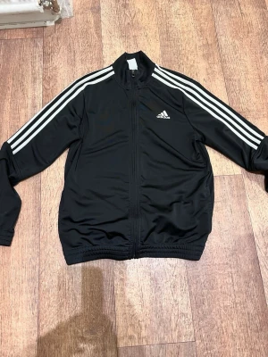 Svart Adidas träningsjacka - Svart träningsjacka från Adidas med klassiska vita ränder längs ärmarna och Adidas-logga på bröstet. Jackan har dragkedja framtill, ståkrage och resår vid ärmslut och midja. Tillverkad i ett glansigt, sportigt material som passar perfekt till träning eller chill.