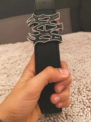 Svart bälte med unik silverspänne - Säljer ett svart bälte i skinn med ett riktigt coolt och unikt silverspänne i grafisk design. Perfekt för att lyfta vilken outfit som helst med en edgy vibe. Bältet har en bred rem och känns riktigt trendigt.