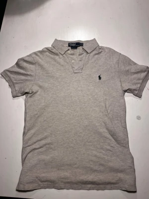 Grå pikétröja från Ralph Lauren - Klassisk beige pikétröja från Ralph Lauren med broderad logga på bröstet. Tröjan har krage, knappar framtill och korta ärmar. Perfekt för en stilren och avslappnad look.