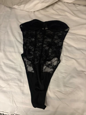 Svart spetsbody utan axelband - Säljer en svart body i spets med strapless design. Bodyn har genomskinliga spetsdetaljer framtill och slät tyg nedtill. Perfekt för dig som gillar en elegant och sexig look. Märke och storlek saknas.