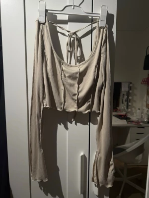 Beige croppad ribbad topp med knyt - Trendig croppad topp i beige med långa ärmar och ribbad struktur. Toppen har snygg knytning i ryggen och vida ärmslut för en extra cool look. Perfekt för dig som gillar unika detaljer och vill sticka ut.