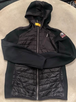 Svart hybridjacka från Parajumpers ”Nolan Boy cardigan” - Snygg svart jacka/cardigan från Parajumpers med huva, quiltade partier fram och bak samt dragkedja. Jackan har en bröstficka med dragkedja och ett broderat märke på ärmen. Perfekt för lager-på-lager och streetwear.  Jackan är i toppskick och passar perfekt vår/höst. Nypris på Kids Brand store 3599.