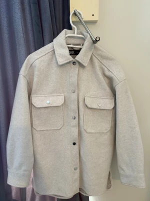  kappa - Stilren ljusgrå overshirt från WAP med två stora bröstfickor och tryckknappar framtill. Overshirten har klassisk krage och är tillverkad i ett mjukt, filtliknande material som ger en clean och modern look. Perfekt lager på lager-plagg för kyligare dagar.