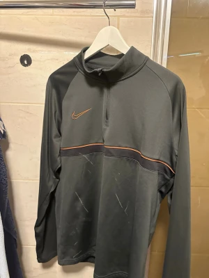 Mörkgrå Nike träningströja herr - Snygg mörkgrå långärmad träningströja från Nike med halv dragkedja och orange detalj på bröstet. Den är lite smutsig men tvätt sker innan köp