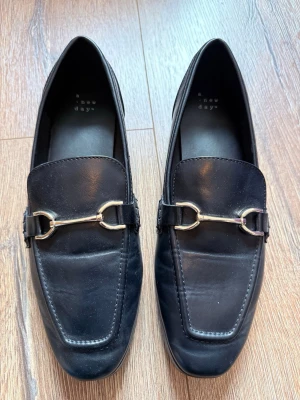 Svarta loafers från A New Day - Stilrena svarta loafers från A New Day med klassisk metallspänne över vristen. Skorna har en fyrkantig tå och är tillverkade i slätt konstläder. Perfekta för en trendig och avslappnad look.  Köpta på Target i USA, storleken är 9 1/2 och passar som ca 40.5-41. Mjuka & sköna att ha på och har används ett par gånger bara. Har en minimal slitning på sulan men inget anmärkningsvärt egentligen