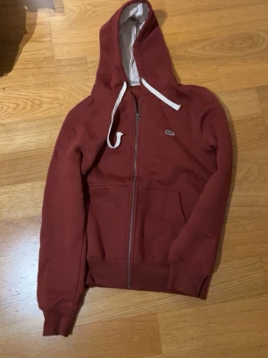 Vinröd hoodie från Lacoste - Säljer en vinröd hoodie från Lacoste med dragkedja och vit dragsko. Klassisk krokodillogga på bröstet och två fickor framtill. Mjuk insida och ribbade muddar vid ärmslut och nederkant. Perfekt för en chill och stilren look.
