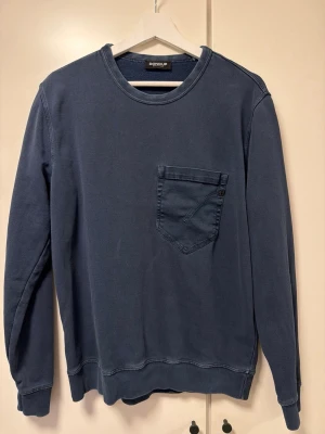 Blå sweatshirt med bröstficka - Snygg blå sweatshirt från Dondup med rund halsringning och en praktisk bröstficka. Tröjan har långa ärmar och ribbade muddar vid ärmslut och nederkant. Perfekt för en avslappnad och stilren look.