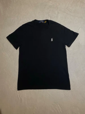 Svart t-shirt från Polo Ralph Lauren - Klassisk svart t-shirt från Polo Ralph Lauren med den ikoniska broderade loggan på bröstet. T-shirten har rund halsringning och korta ärmar. Tillverkad i mjuk bomull för en skön känsla och enkel stil.