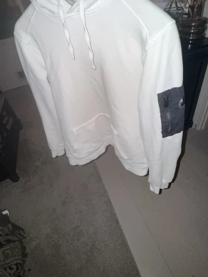 Vit hoodie från Snoot med ärmficka - Vit hoodie från Snoot med klassisk huva och känguruficka framtill. På vänster ärm finns en svart ficka med dragkedja och Snoot-logga. Hoodien har en avslappnad passform och är tillverkad i mjukt material, perfekt för en chill och stilren look.