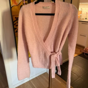 Rosa omlottkofta från Gina Tricot - Mysig rosa stickad omlottkofta från Gina Tricot med knytband i midjan och långa ärmar. Koftan har en djup v-ringning och är tillverkad i ett fluffigt, mjukt material som känns extra skönt mot huden.