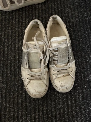 Vita Valentino Garavani sneakers - Snygga vita sneakers från Valentino Garavani med klassisk låg siluett. Skorna har vita snören, detaljer i grått och en bred plös med logotyp. Tillverkade i läder med gummisula och diskreta nitar bak på hälen. Perfekta för dig som gillar exklusiva märken. Accepterat skick mestadels ytligt smuts som går att städa bort. Slitna vid hälarna varav priset. Inköps pris cirka 7000