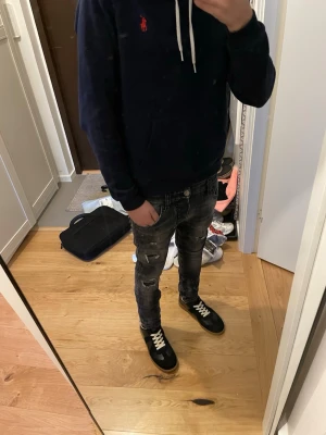 Svarta slitna jeans från Dsquared2 - Säljer ett par svarta jeans från Dsquared2 med slitningar och färgstänk för en edgy look. Jeansen har smal passform och klassisk femficksdesign. Medföljer en vit bälte med stor metallspänne som ger extra attityd till outfiten. Bältet kommer inte med. Jag möts bara upp om man ska köpa.