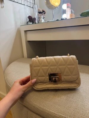 Beige quiltad axelväska med kedja - Säljer en beige quiltad axelväska med guldfärgad kedjerem och snyggt rutmönster. Väskan har ett metallspänne framtill och är tillverkad i konstläder. Perfekt storlek för det viktigaste och ger en trendig touch till din outfit.