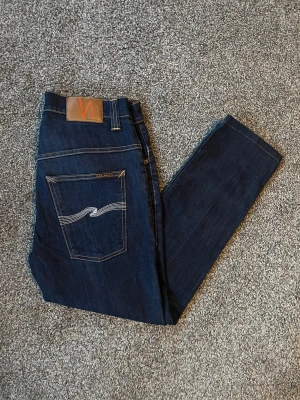 Mörkblå jeans från Nudie Jeans - Säljer ett par mörkblå jeans från Nudie Jeans med klassisk femficksmodell och kontrastsömmar. Jeansen har rak passform och en snygg broderad detalj på bakfickan. Tillverkade i denim med en stilren look.
