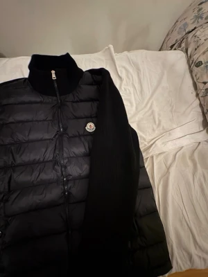 Svart cardigan Moncler - Snygg svart dunväst från Moncler med quiltad design och hög krage. Västen har dragkedja framtill och ett Moncler-märke på bröstet. Perfekt för lager-på-lager och håller dig varm med sin dunfyllning.