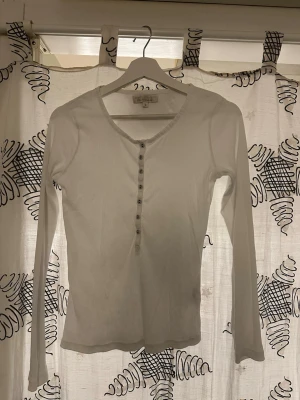 Vit långärmad topp Capri Collection - Säljer en enkel och stilren vit långärmad topp från Capri Collection. Toppen har v-ringning och knappar framtill, vilket ger en snygg detalj. Tillverkad i ett mjukt ribbat material som sitter skönt på kroppen. Perfekt att matcha med jeans eller kjol.