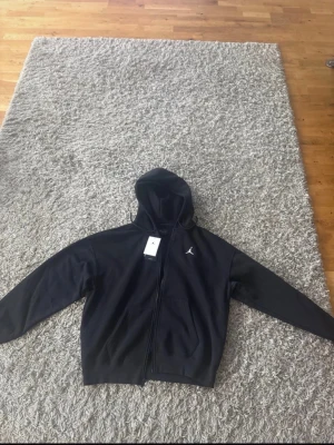 Svart Jordan hoodie med dragkedja - Snygg svart hoodie från Jordan med dragkedja framtill och klassisk huva. Ikoniska Jumpman-loggan i vitt på bröstet. Hoodien har två fickor fram och ribbade muddar vid ärmar och nederkant. Perfekt för en sportig och avslappnad stil.