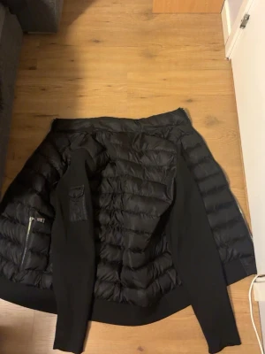 Svart dunväst från Moncler - Snygg svart dunväst från Moncler med quiltad design och stickade detaljer på sidor och ärmar. Västen har dragkedja, fickor och en Moncler-logga på ärmen. Perfekt för lager-på-lager och streetwear-vibe.