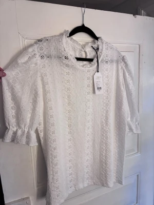 Vit spetsblus från Jyothi - Superfin vit blus från Jyothi i spets med romantiska volangdetaljer vid ärmslut och hals. Blusen har trekvartsärm och ett diskret hålmönster som ger en elegant look. Perfekt att styla med jeans eller kjol för en trendig outfit. Helt ny med prislapp kvar.