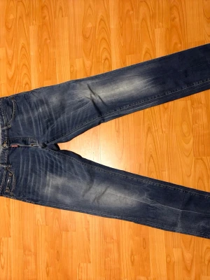 Blå Dsquared Jeans - Säljer ett par klassiska blå jeans från Dsquared med raka ben och snygga slitningar. Jeansen har fem fickor, knappgylf och den ikoniska röda Dsquared lappen vid fickan. Tillverkade i slitstark denim som passar perfekt till en avslappnad stil.
