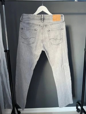 Grå raka jeans från Jack & Jones - Säljer ett par ljusgrå jeans från Jack & Jones med rak passform och klassisk femficksdesign. Jeansen har en snygg tvättad look och är tillverkade i bomull. Perfekta för en avslappnad och trendig stil.