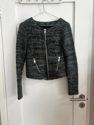 Gråmelerad cardiganjacka med dragkedja - Snygg gråmelerad cardiganjacka med raka linjer och silverfärgade dragkedjor framtill. Jackan har två fickor med dragkedja och är tillverkad i ett mjukt, strukturerat tyg. Långärmad och något kortare modell som passar perfekt till jeans eller kjol.