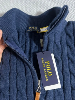 Blå kabelstickad tröja Polo Ralph Lauren - Mörkblå kabelstickad tröja från Polo Ralph Lauren med halv dragkedja och ribbade muddar. Tröjan har det klassiska röda Polo-logot på bröstet och är tillverkad i mjukt material. Perfekt för en stilren och avslappnad look.