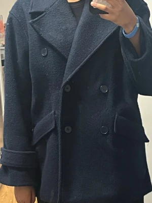 Mörkblå dubbelknäppt ullrock - Snygg mörkblå rock med dubbelknäppning och breda slag. Rocken har två stora fickor framtill och uppvikta ärmslut med knappdetalj. Tillverkad i ett ulligt material som håller dig varm och stilren under kalla dagar.