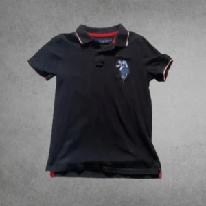 Polo piké - Svart polo piké från U.S. Polo Assn med klassisk krage och korta ärmar. Röd och vit kant på krage och ärmslut, stor röd siffra 3 på ena ärmen och broderad polospelare på bröstet. Tillverkad i mjuk bomull, perfekt för en sportig och chill stil.