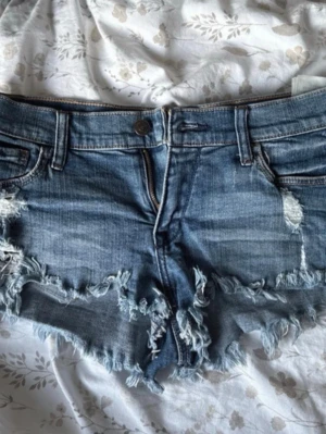 Lågmidjade jeansshorts  - Snygga blå jeansshorts från Abercrombie & Fitch med slitna detaljer och fransig kant. Klassisk femficksmodell med låg midja och råa kanter för en avslappnad look. Perfekta för varma dagar och enkel att matcha med allt.