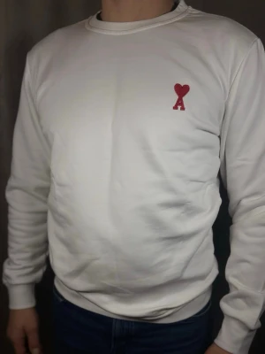 Vit sweatshirt från Ami Paris - Säljer en stilren vit sweatshirt från Ami Paris med det ikoniska röda Ami de Coeur-emblemet broderat på bröstet. Tröjan har rund halsringning, ribbade muddar och är tillverkad i mjukt bomullsmaterial. Perfekt för en clean och trendig look.