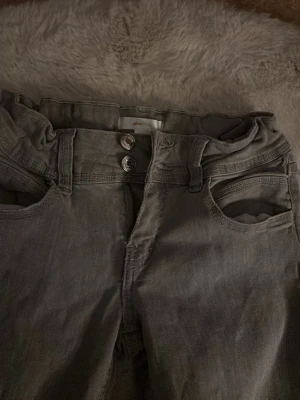 Gråa bootcut jeans med mid waist - Säljer ett par gråa bootcut jeans med hög midja och dubbla knappar i linningen. Jeansen har klassiska fem fickor och är tillverkade i mjukt denimtyg. Perfekta för dig som gillar en avslappnad men trendig look.