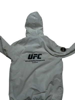 Vit UFC vindjacka med huva - Snygg vit vindjacka från UFC med svart dragkedja och huva. Stor UFC-logga på ryggen och patch på ena ärmen. Lätt material, perfekt för blåsiga dagar. Sportig och stilren design som sticker ut.(köpt i Paris hittar ingenstans på internet)